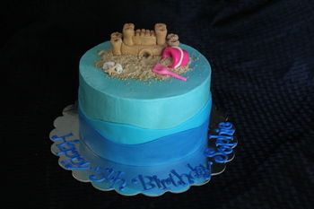 Sandcastle and sea shells with bucket and shovel in the sand.  8" round 4 layer cake covered in buttercream with marshmallow fondant waves and hand sculpted decorations.  Sand is cookie crumbs.